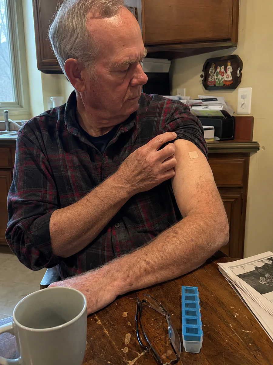 A man in his sixties at a kitchen table with his sleeve rolled up, looking at a small adhesive patch on his upper arm.